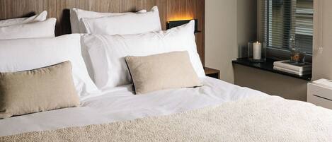 A neatly made bed with white linens and multiple pillows, a wooden headboard, a bedside table with a lamp, and a window with blinds.