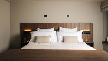 Cama bem arrumada com cabeceira de madeira, duas mesas de cabeceira com luminárias e uma pequena estante com objetos decorativos.