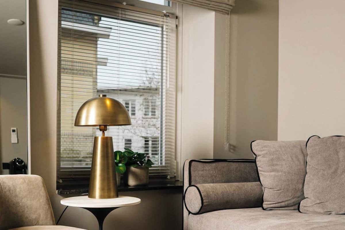 A modern living room with a round armchair, a sofa with cushions, a small round table with a lamp, and a window with blinds.