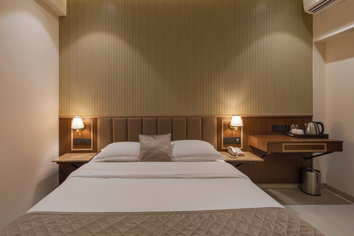 Ein Hotelzimmer mit einem großen Bett, zwei Nachttischen mit Lampen, einem Holznachttisch, einem kleinen Tisch mit Wasserkocher und einem Papierkorb.