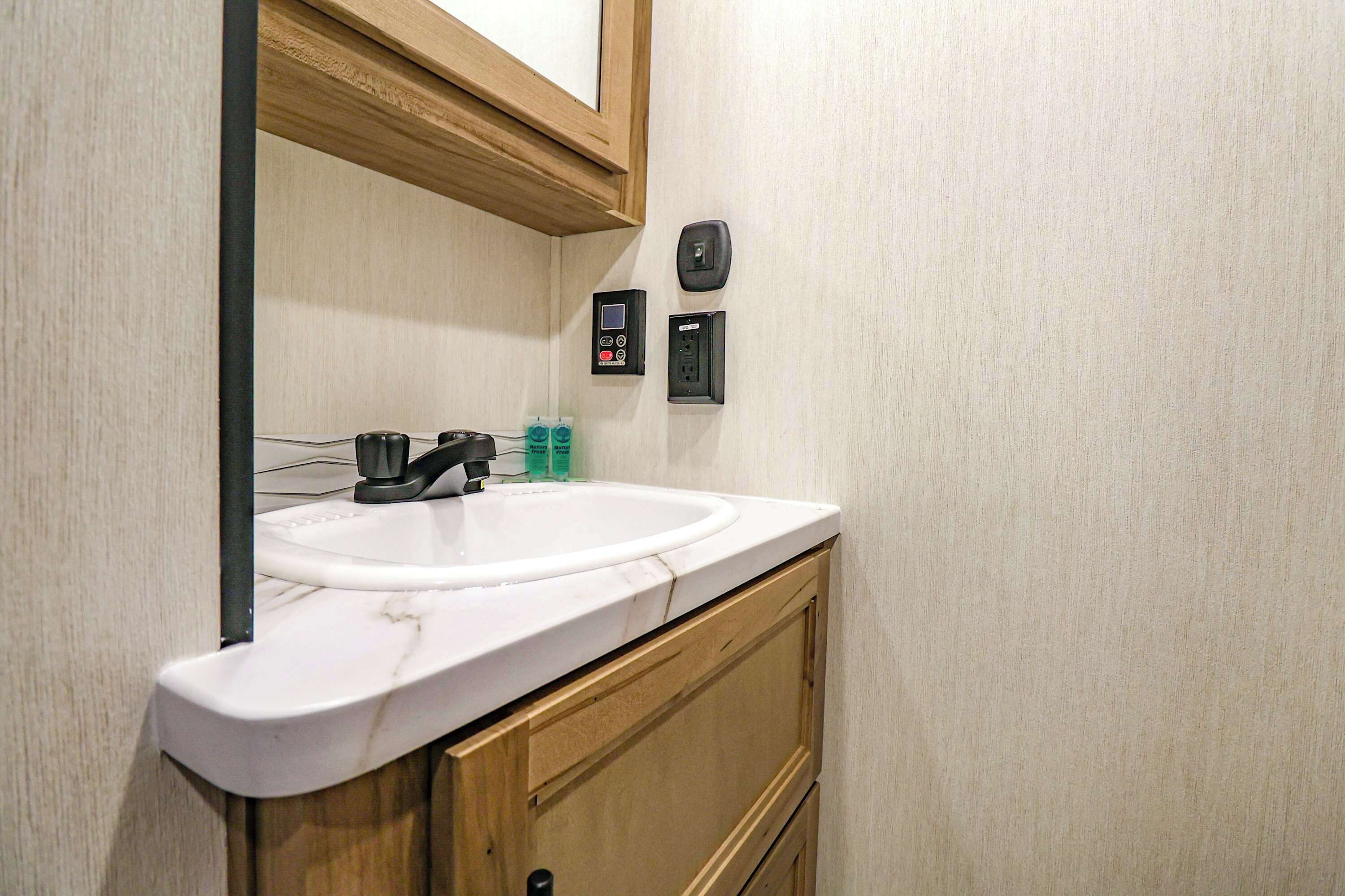 Un baño con un lavabo blanco, armarios de madera, un espejo y dos enchufes en la pared.