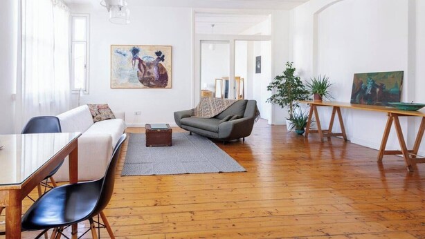 A spacious living room with a wooden floor, a grey sofa, a dining table with chairs, and a painting on the wall.