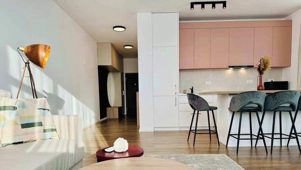 A modern living room with a round wooden coffee table, a sofa, a floor lamp, and a kitchen area with pink cabinets and two bar stools. - Joy City Stay Nord One N2-1 (Timișoara)