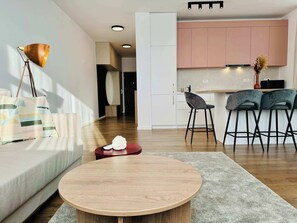 Un soggiorno moderno con un tavolino rotondo in legno, un divano, una lampada da terra e un angolo cottura con pensili rosa e due sgabelli da bar.