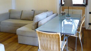 Un salon moderne avec une table à manger au plateau en verre, un canapé d’angle beige et un fauteuil en osier.
