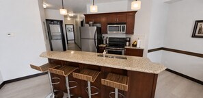 A kitchen with a granite countertop, stainless steel appliances, wooden bar stools, and pendant lighting. - Alpine Splash Getaway – Pool, Hot Tub & Waterslide! (Canmore)