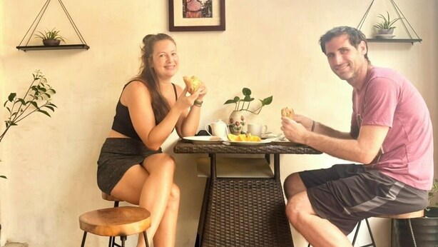 A dining area with a wicker table, two chairs, and a window with a view. There are framed pictures on the wall and small potted plants on shelves. - Good Morning Hoi An Homestay and Hostel (Da Nang)