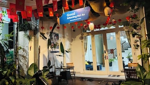 A cozy indoor space with a sign reading "Good Morning Hong An Homestay & Hostel," featuring a dining area with chairs and tables, potted plants, and a scooter parked nearby. - Good Morning Hoi An Homestay and Hostel (Da Nang)