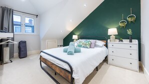 Ein Schlafzimmer mit schrägem Dach, einem Bett mit weißer Bettwäsche, einem Schminktisch, einer Nachttischlampe und einem Fenster mit Vorhängen.