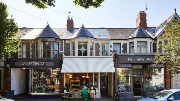 Eine Reihe von Geschäften mit den Schildern 'AL PONTE' und 'The Frame Shop', ein Café mit Außenplätzen und abgestellte Autos.
