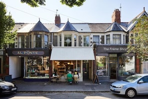 Una hilera de tiendas, con los letreros 'AL PONTE' y 'The Frame Shop', un café con terraza y coches estacionados.