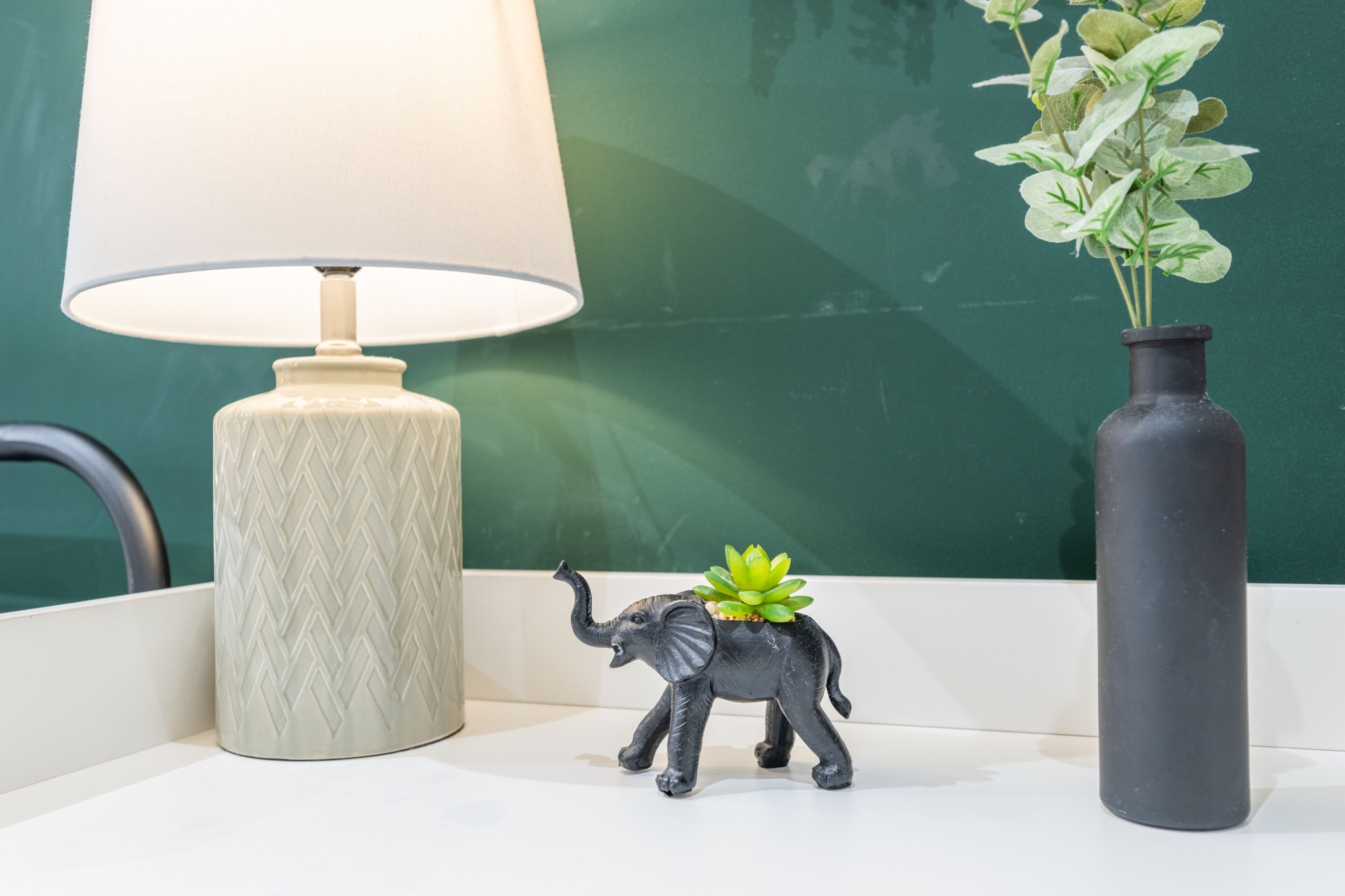 Un bureau avec une lampe, une petite figurine d’éléphant accompagnée d’une plante, et un vase avec des feuilles vertes.