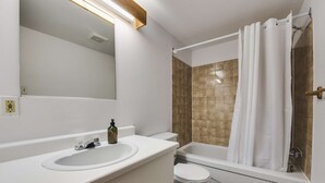 Un baño con lavamanos, inodoro y bañera blancos. Hay un espejo sobre el lavamanos y una cortina de ducha sobre la bañera.