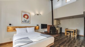 Un dormitorio con una cama grande, cabecera de madera, televisor montado, chimenea, una mesa de comedor con sillas y un ventilador de techo.