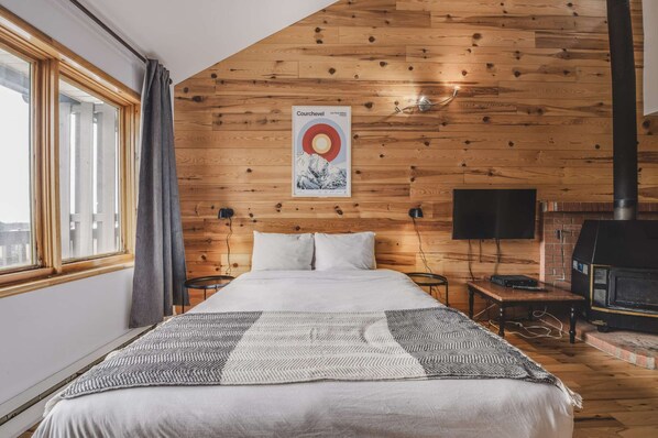 Une chambre avec un plafond en lambris de bois, un lit recouvert d’une couverture à motifs, une affiche accrochée au mur, un téléviseur et une cheminée.