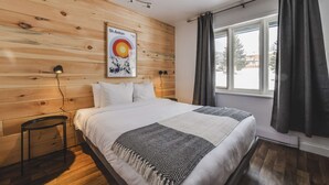 A bedroom with a wooden wall, a bed with white bedding, a black and white throw, a small table, and a window with curtains. - Studio St-Anton 4 (1 Q) - 2 people (Saint-Ferréol-les-Neiges)