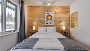 Habitación de hotel con pared de paneles de madera, cama con colcha estampada, dos lámparas de noche, un cartel en la pared y ventana con vistas a árboles y un coche.