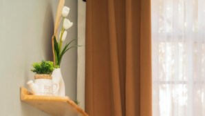 A neatly made bed with white bedding and two pillows, a wooden shelf with a small plant and a decorative item, and a window with sheer curtains.