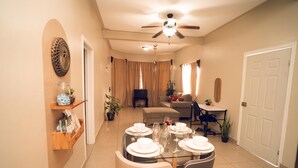 A dining area with a glass table set for four, surrounded by chairs. There is a ceiling fan above, and a television is visible in the background.
