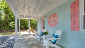 A covered porch with white chairs, a table, and a wall with decorative cutouts. - Experience coastal bliss at Sand Dollar Daze, a spacious 3-bedroom home in Oak Island. (Oak Island)