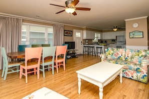 Uma sala de estar com uma mesa de jantar, cadeiras coloridas, uma mesa de centro branca, um sofá floral, um ventilador de teto e uma área de cozinha com banquetas.