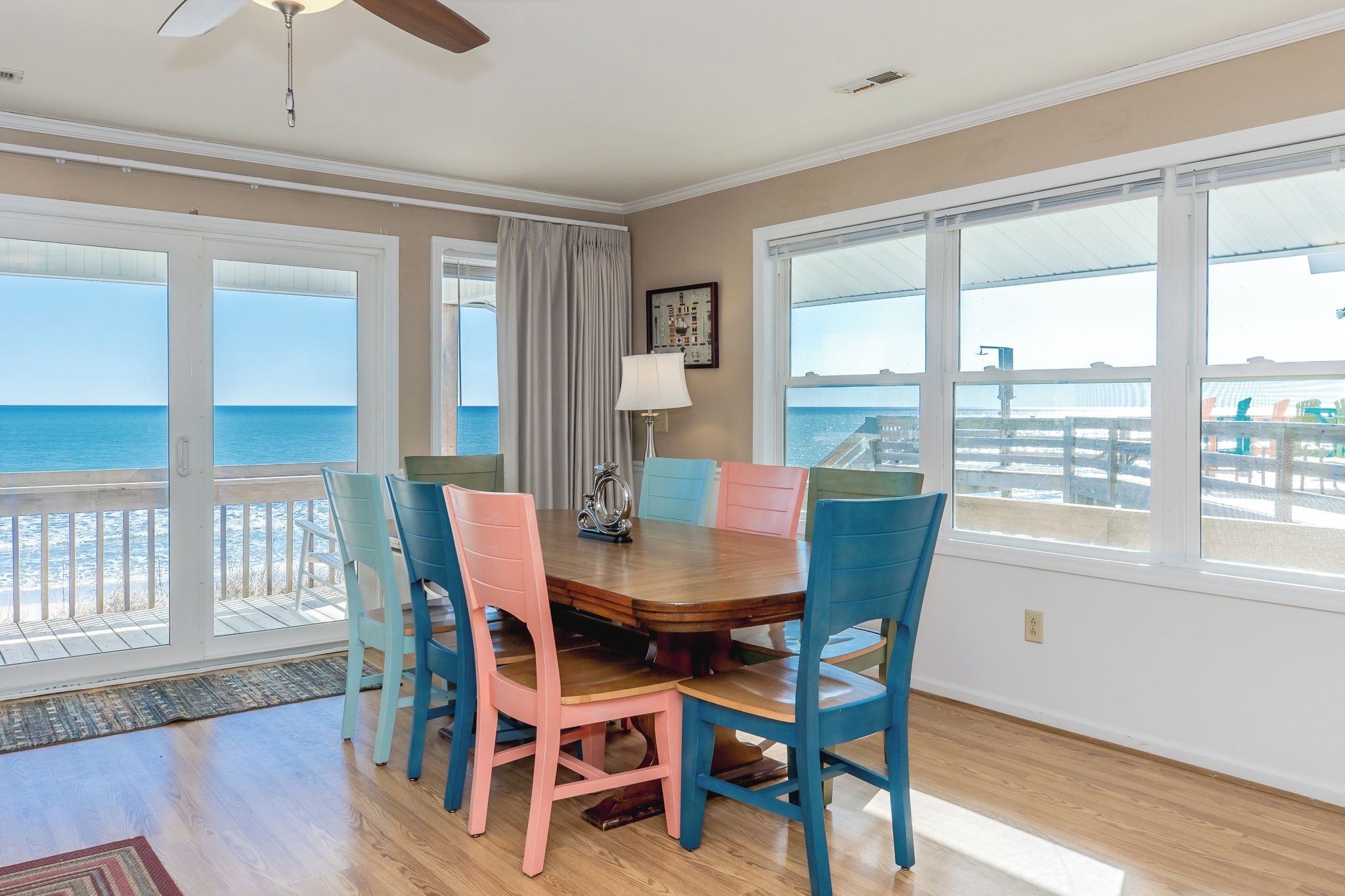 A dining area with a wooden table and colorful chairs, large windows with a view of the ocean, and a ceiling fan.