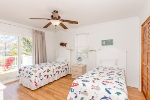 Quarto com duas camas, ventilador de teto, janela com persianas e vista para o exterior.
