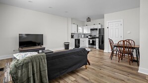 A modern living room with a flat-screen TV, a dark sofa, and a dining area with wooden chairs and a table.