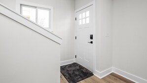 A clean, white hallway with a wooden floor, a black rug, a white door with a window, and a staircase leading to an upper level.