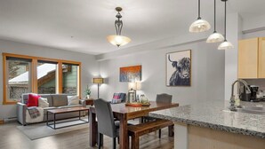 Eine moderne Küche mit Granit-Arbeitsplatten, ein Essbereich mit Holztisch und -bänken, ein Wohnzimmer mit Sofa und Kamin, und ein großes Fenster mit Blick auf eine verschneite Landschaft.