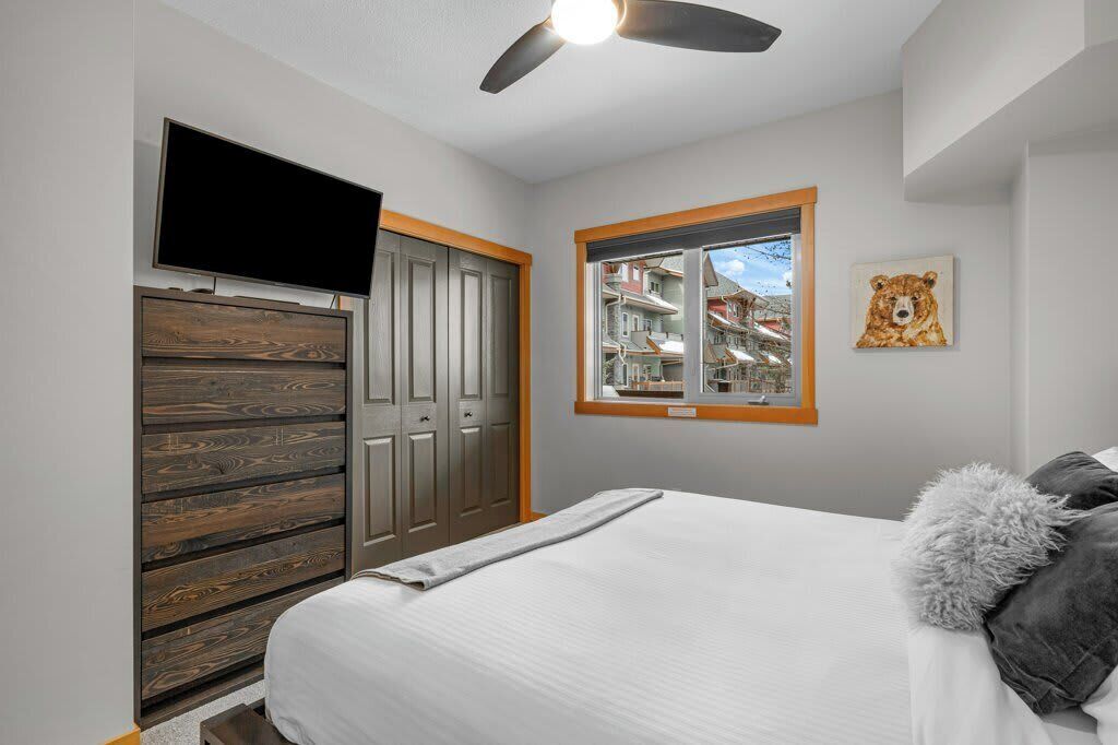 A bedroom with a bed, a TV mounted on a wooden cabinet, a window with a view, and a ceiling fan.
