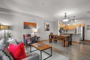 Ein modernes Wohnzimmer mit einer Couch, einem Couchtisch und einem Essbereich mit einem Holztisch und Stühlen. Im Hintergrund ist eine Küche mit Holzausbau und Edelstahlgeräten zu sehen.