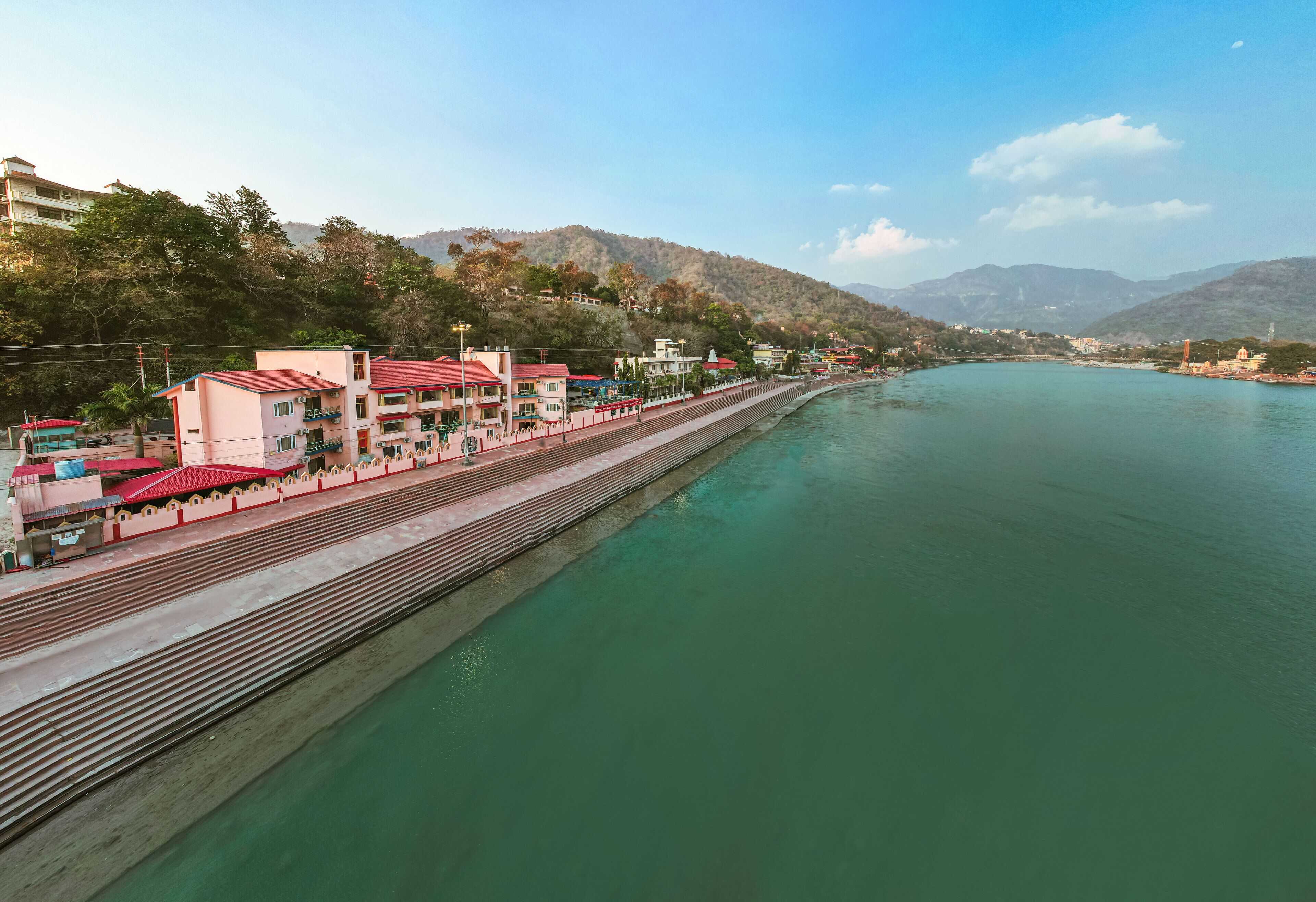 A riverside promenade with buildings, a bridge, and a mountainous backdrop.