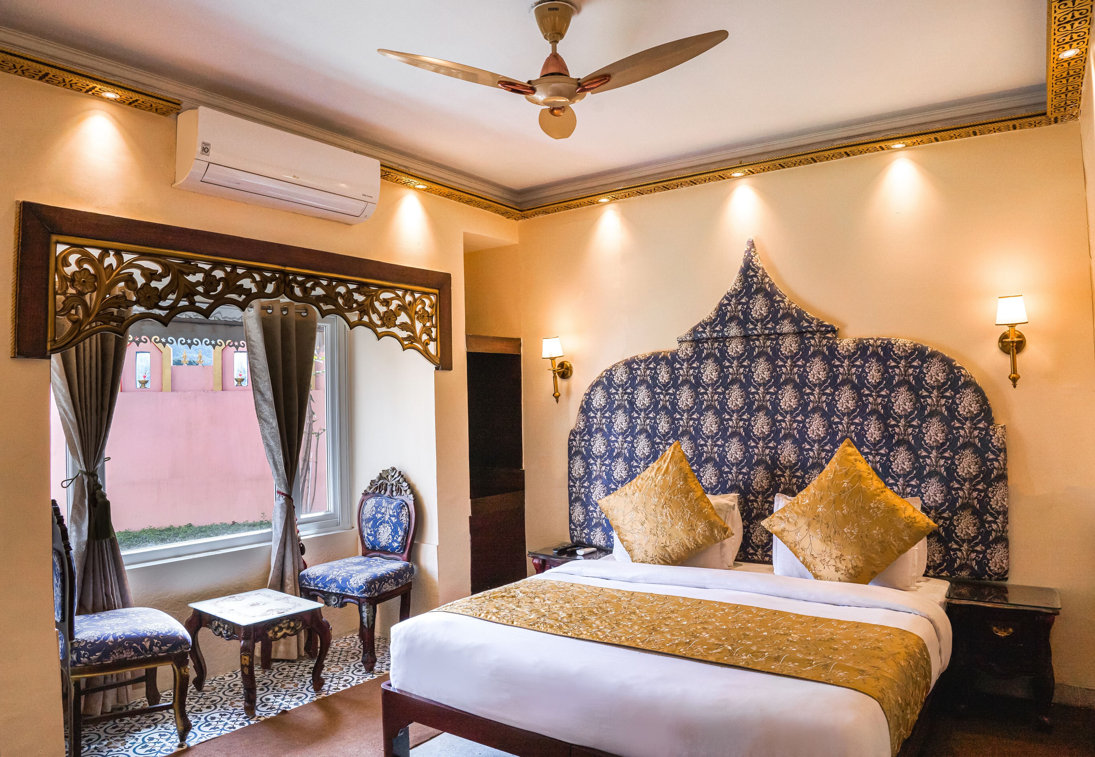 Quarto de hotel com uma cama grande, um ventilador de teto, cabeceira decorativa, duas cadeiras, uma mesa pequena e uma janela com cortinas.