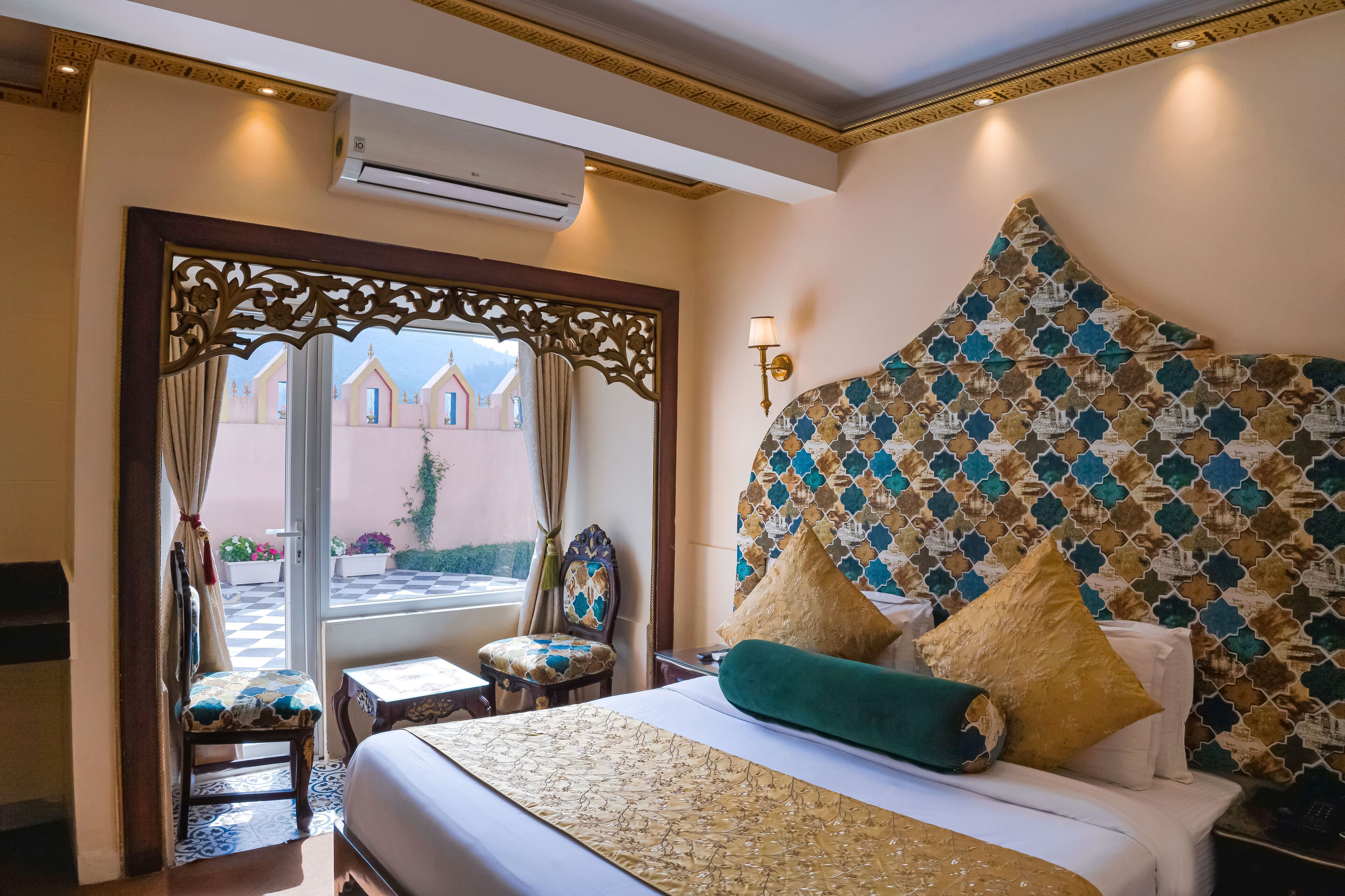 A hotel room with a large bed, a decorative headboard, a small table, and a chair. There is a window with a view of buildings outside.