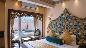 A hotel room with a large bed, a decorative headboard, a small table, and a chair. There is a window with a view of buildings outside.