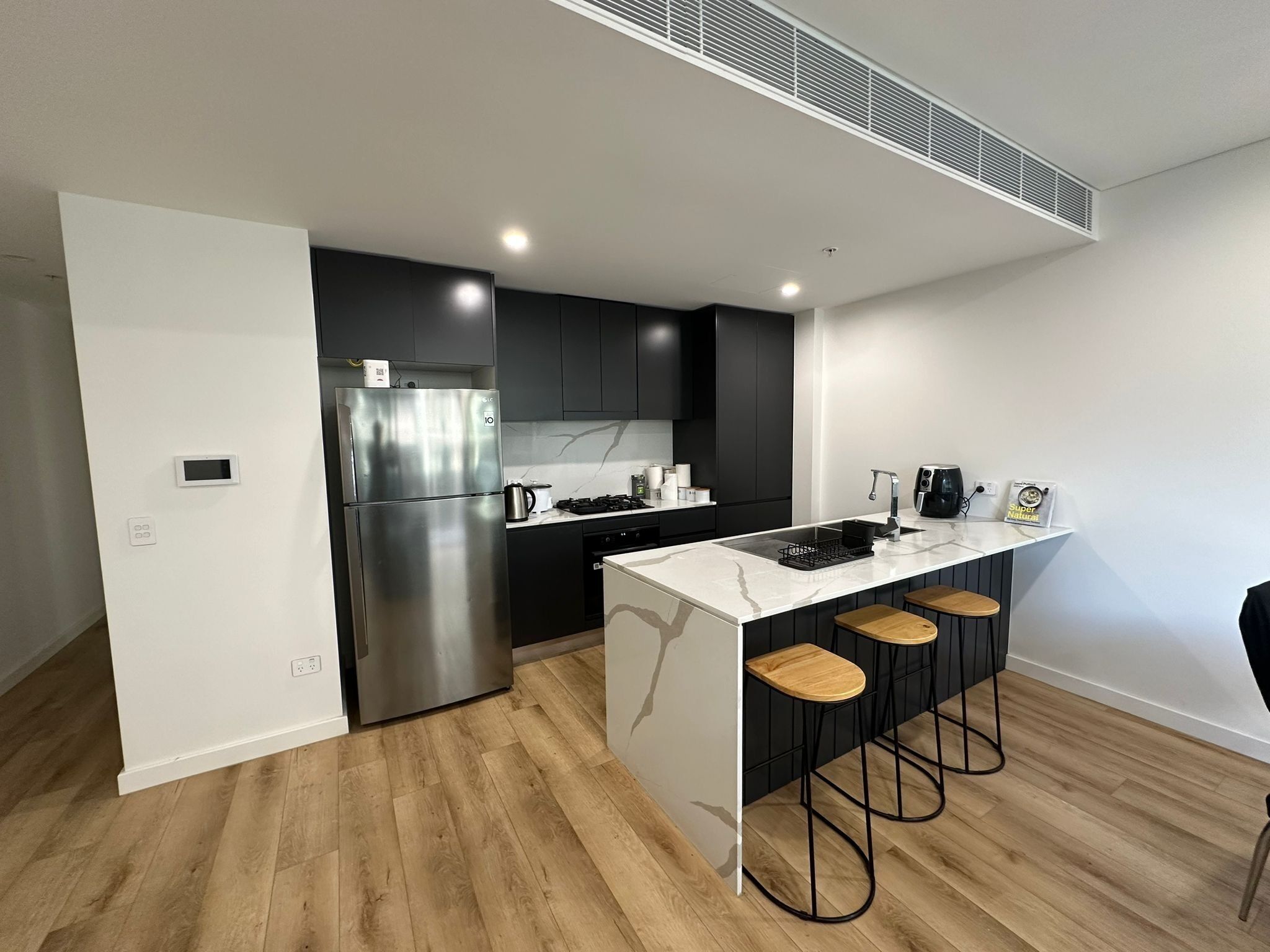 A modern kitchen with a stainless steel refrigerator, a built-in oven, and a marble countertop. There are three wooden stools at the island.