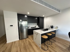 A modern kitchen with a stainless steel refrigerator, a built-in oven, and a marble countertop. There are three wooden stools at the island. - Sydney"Modern 2-Bedroom Apartment near Airport & Beach – Rockdale train  NSW (Rockdale)