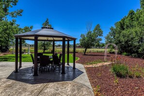 A gazebo with a table and chairs, surrounded by a landscaped garden with trees and a clear sky.