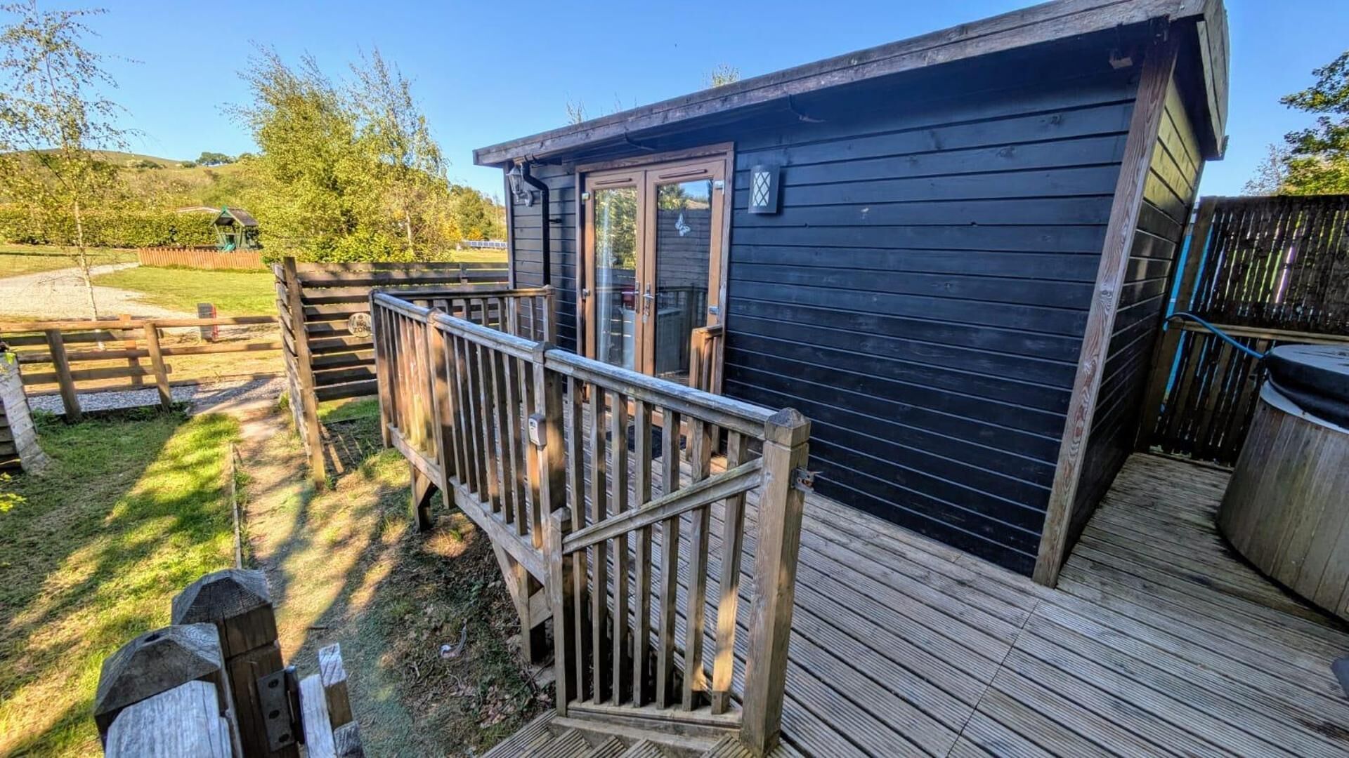 A wooden deck with a metal railing, a small building with a dark exterior, and a fenced area with trees.