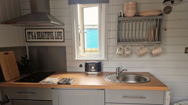 A compact kitchen with a wooden countertop, a built-in oven, a stainless steel sink, and a window with a view of a blue building. - Glan yr Afon Un - Relax, Recuperate, Enjoy (Welshpool)