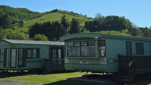 A row of green mobile homes with a grassy hill in the background. - Glan yr Afon Un - Relax, Recuperate, Enjoy (Welshpool)