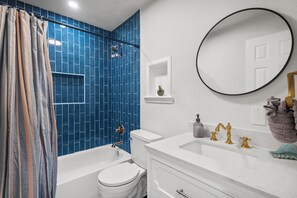 Ein Badezimmer mit blauen Fliesen, einer weißen Badewanne, einer weißen Toilette, einem weißen Waschbecken mit goldenen Armaturen und einem runden Spiegel.