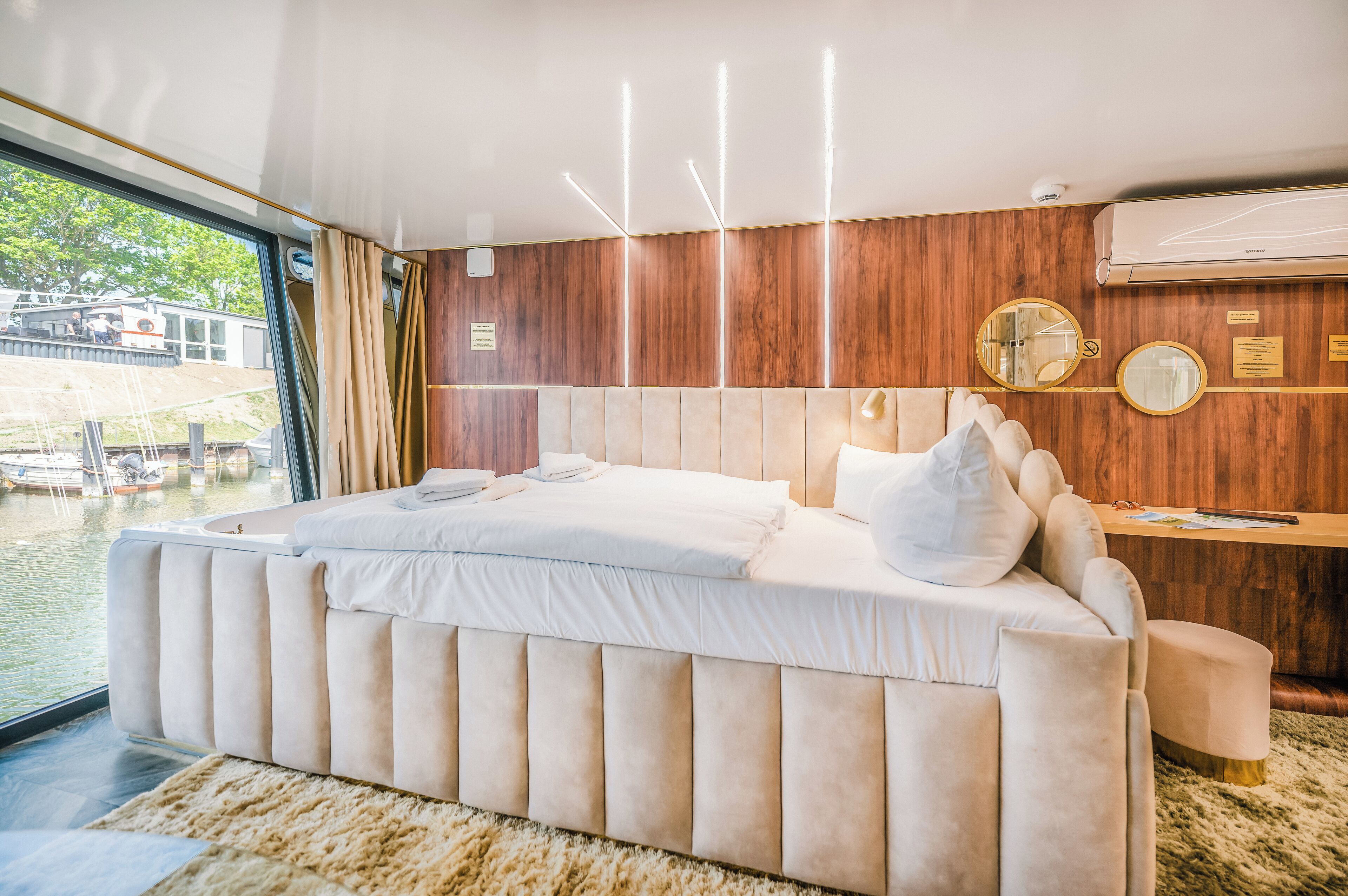 A modern bedroom with a large bed, wooden panel walls, and a view of boats through a large window.