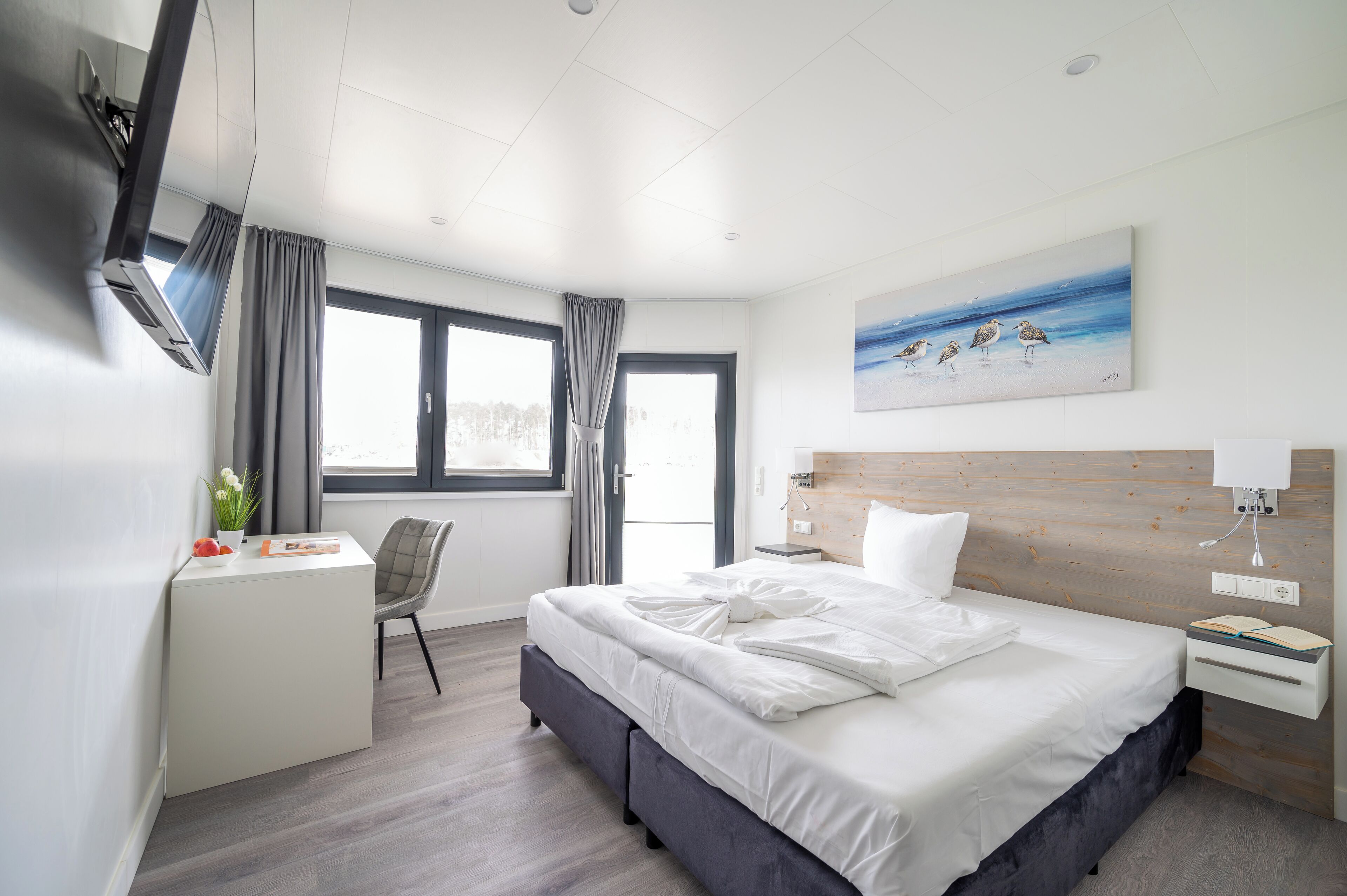 A modern hotel room with a large bed, a desk, a chair, and a painting of birds on the wall.