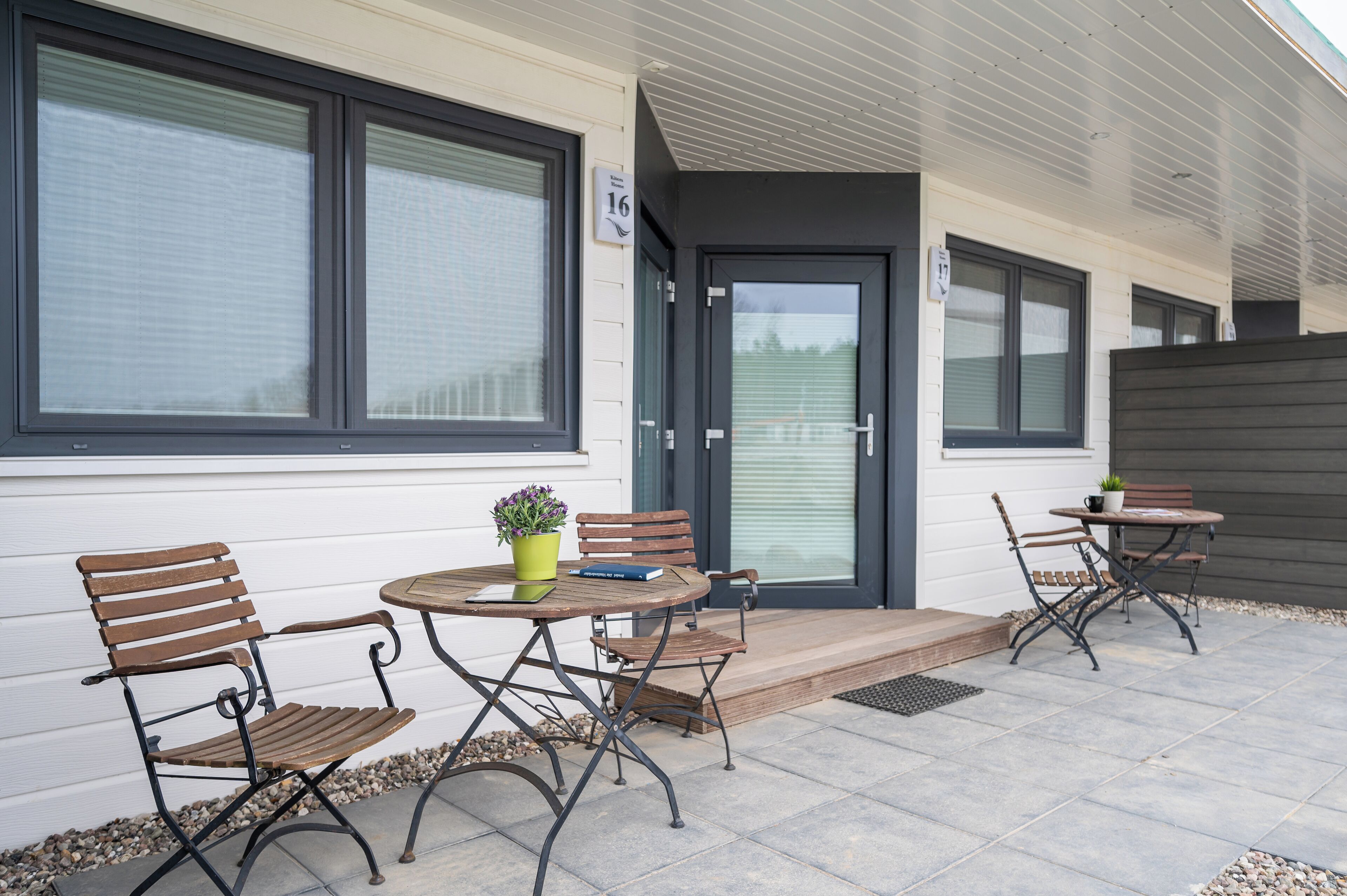 An outdoor patio with two tables and four chairs, a small potted plant, and a door with a number sign.