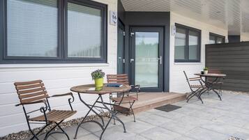 Une terrasse extérieure avec deux tables et quatre chaises, une petite plante en pot et une porte portant un numéro.