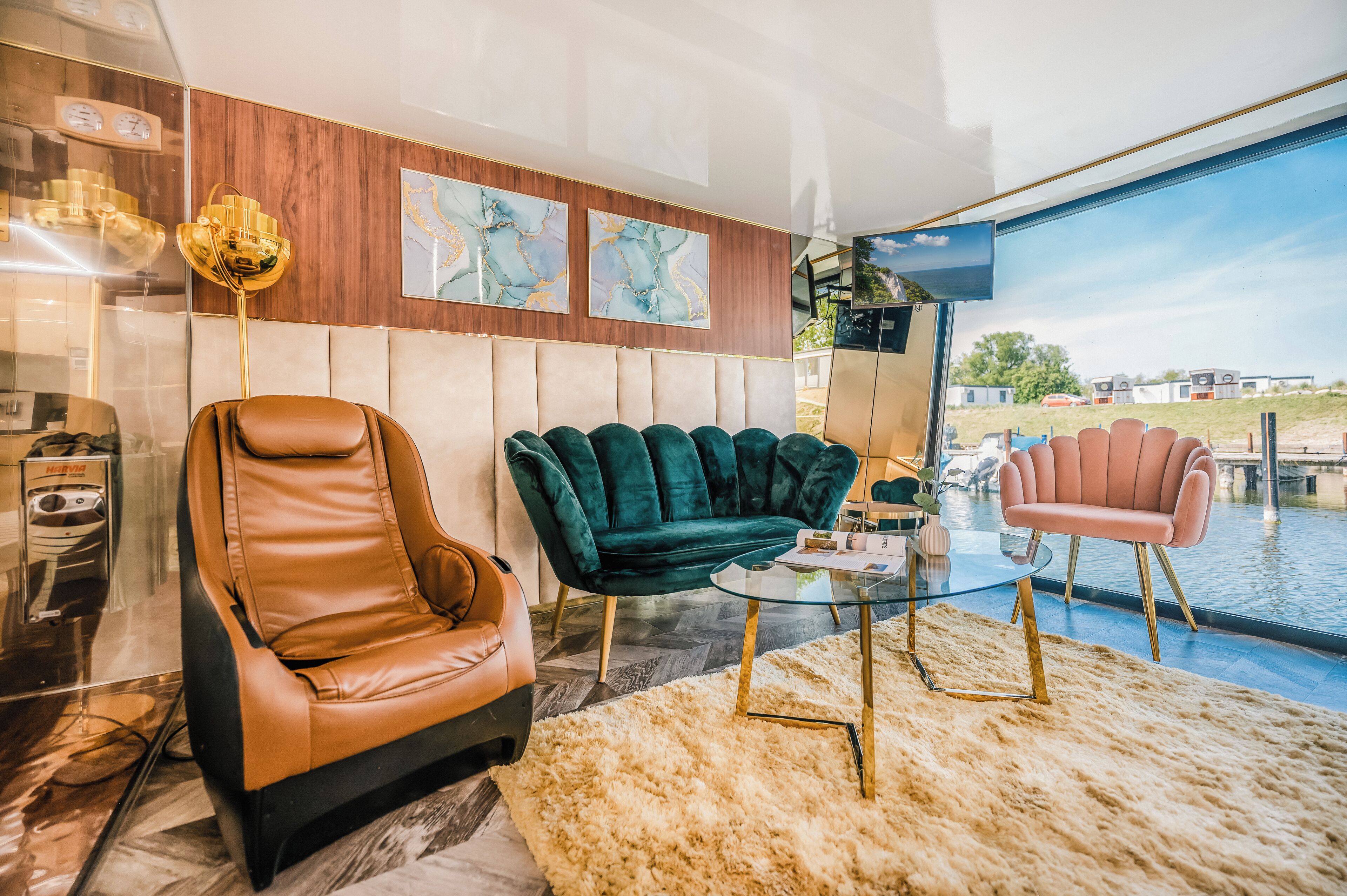 A modern living room with a leather armchair, a green velvet sofa, and a pink chair. There is a glass coffee table and a large window with a view of a body of water and buildings.