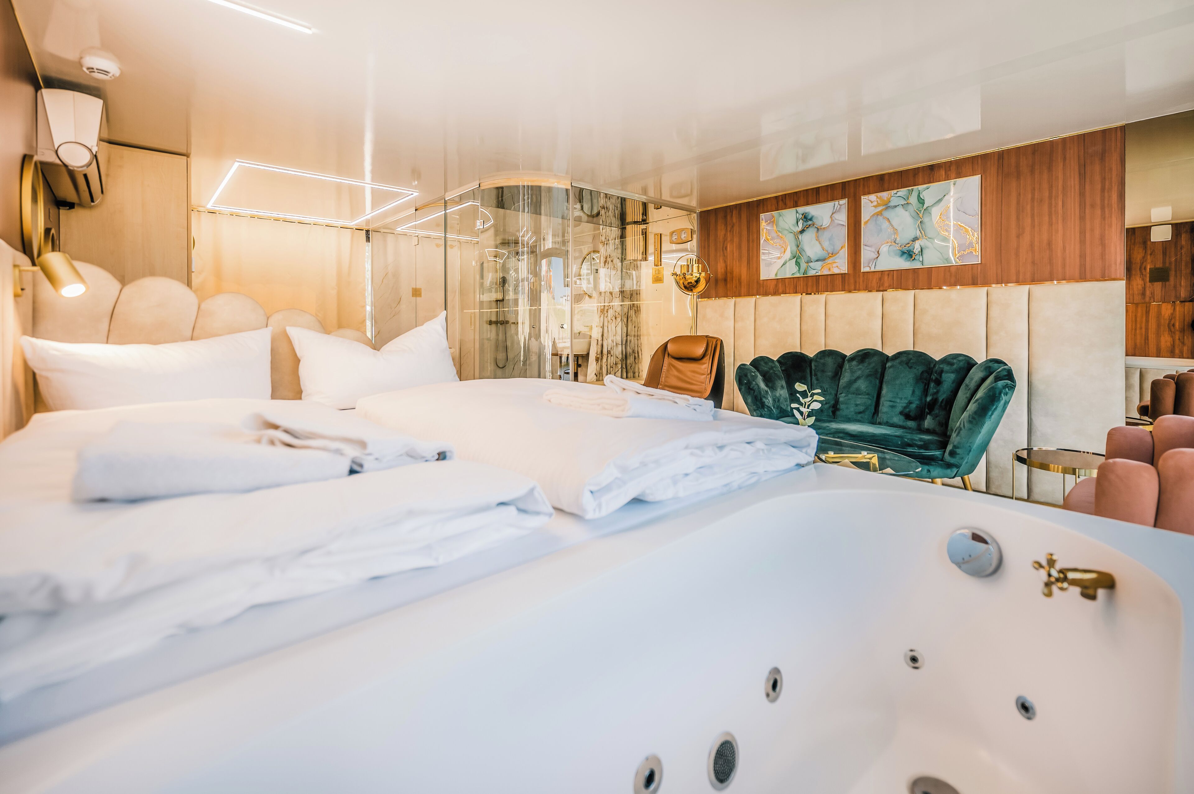 A modern hotel room with a large bed, a sitting area with a green velvet chair, a bathtub, and a wooden panel wall.