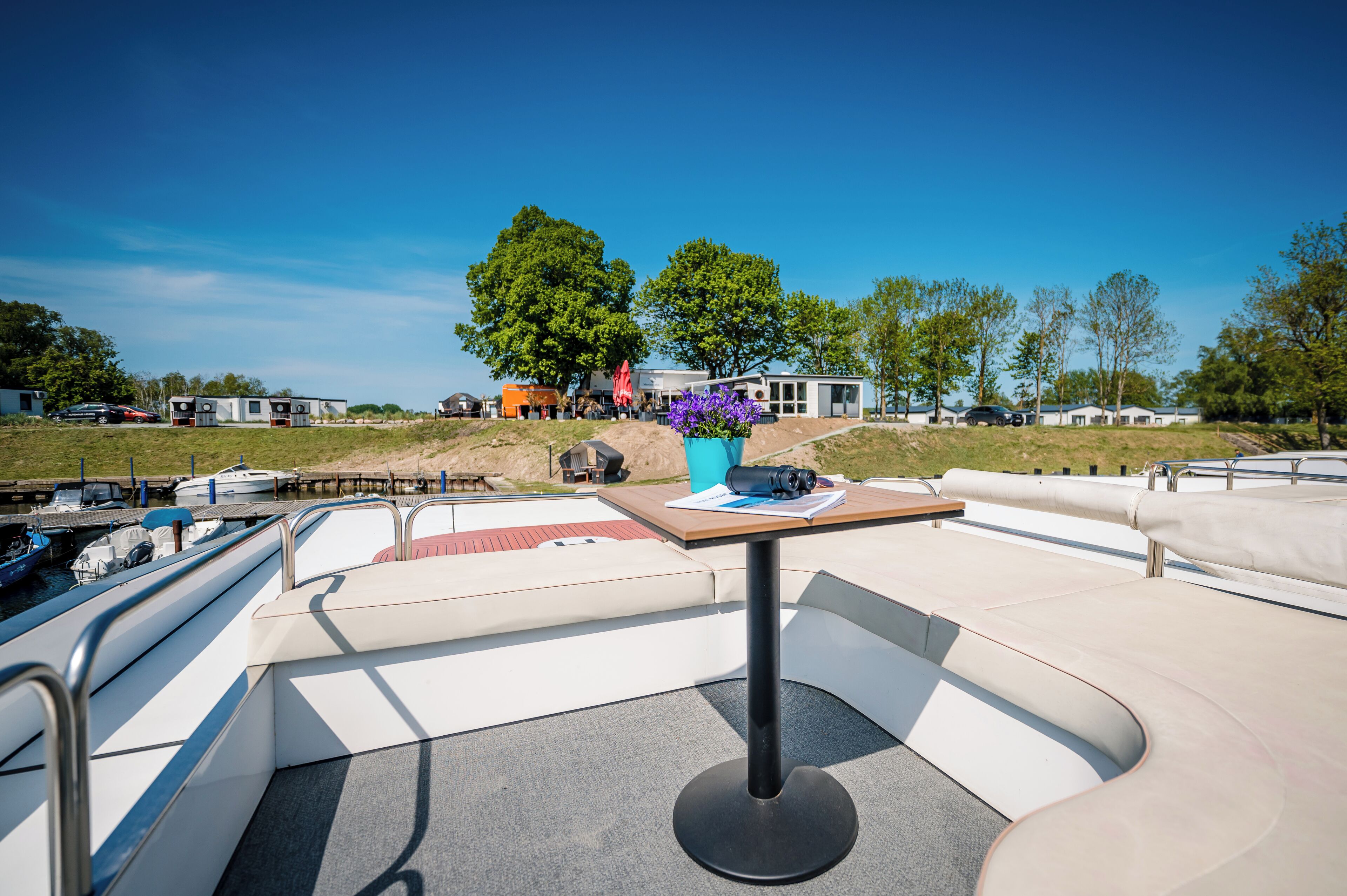 Un pont de bateau avec des sièges, une table sur laquelle est posé un appareil photo, et une vue sur la marina et les bâtiments qui se dressent à l’arrière-plan.